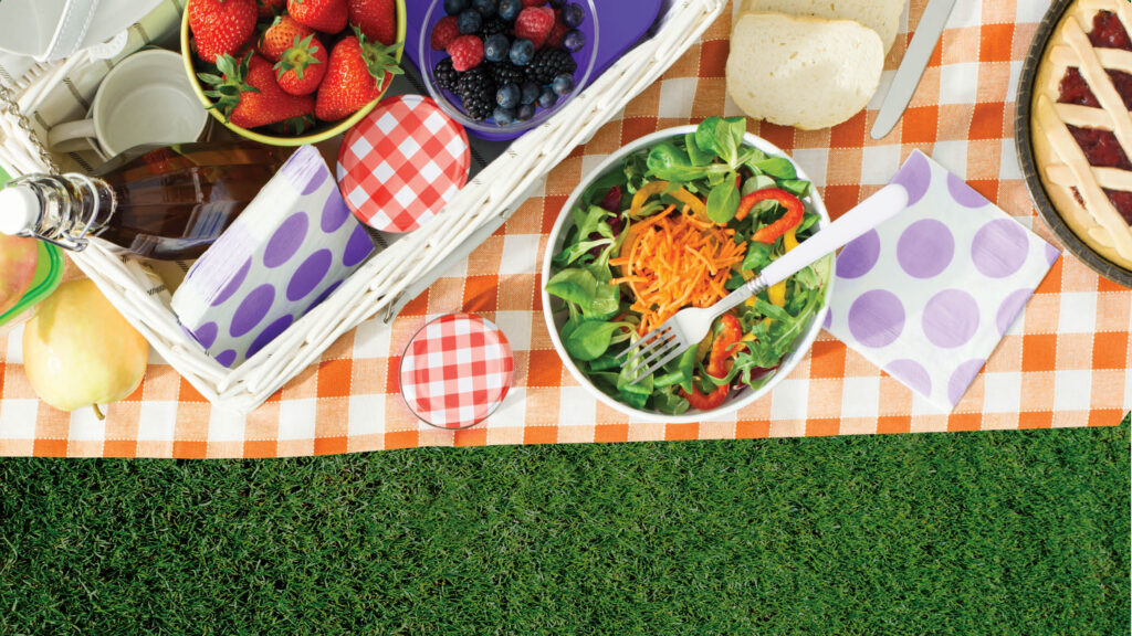 picture of a blanket on grass with a basket of fruits and bowl of salad, pie, and bread