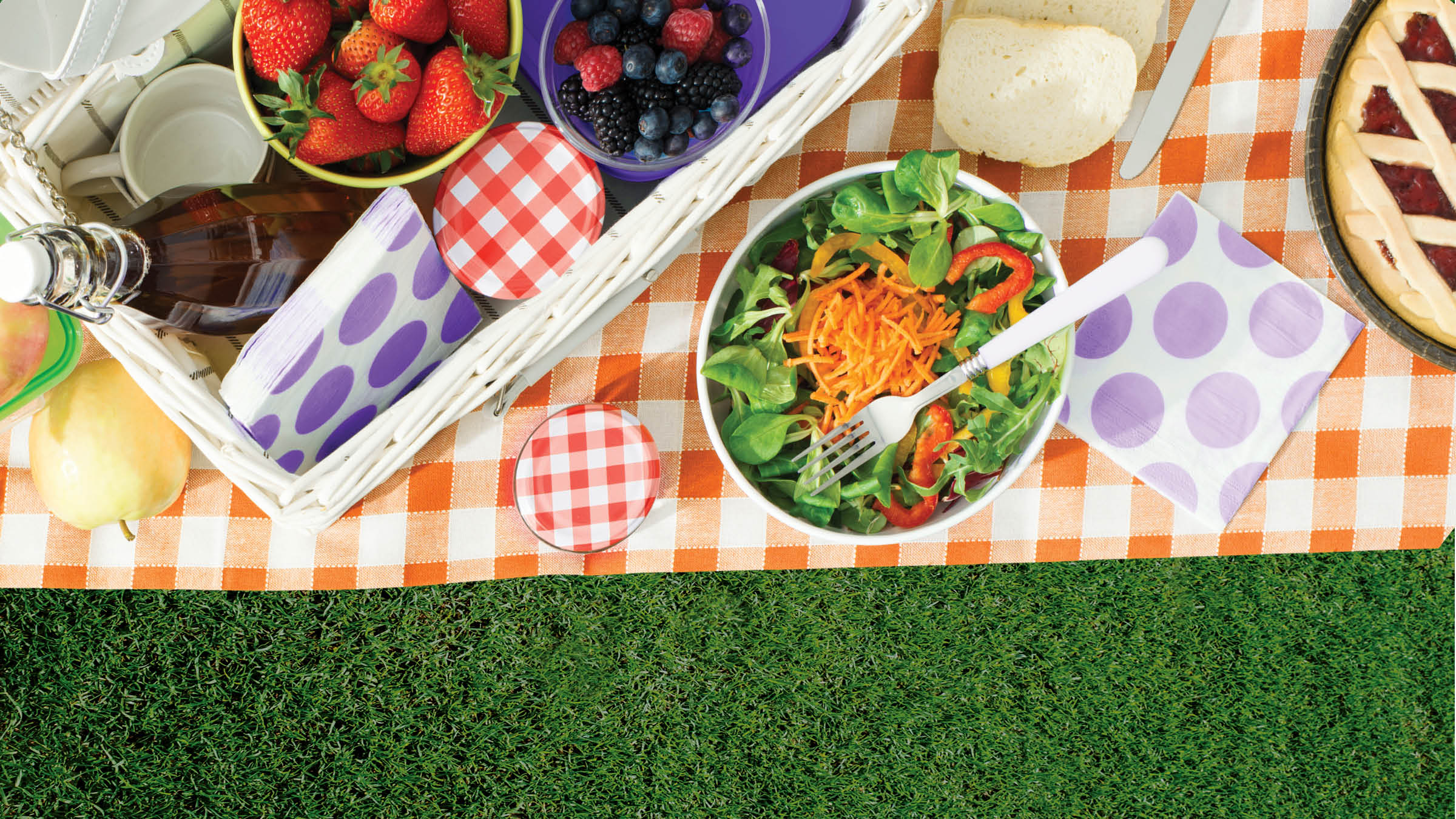 picture of a blanket on grass with a basket of fruits and bowl of salad, pie, and bread